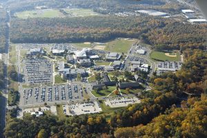 This color photograph shows an aerial view of Camden County College's buildings and sports fields. The parking lots are nearly filled with cars and the surrounding trees showcase autumn colors.
