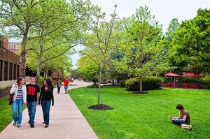 This color photograph shows the quad area of Rutgers University - Camden. Several students walk on the sidewalk or sit on the grass.