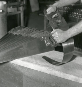 This black and white photo shows a factory worker bending a metal saw blade to ensure its sturdiness before shipment.