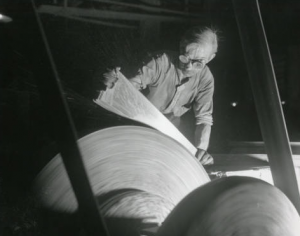 This black and white photograph shows a factory worker wearing goggles and pressing a metal saw blade against a spinning wheel to smooth out one side.