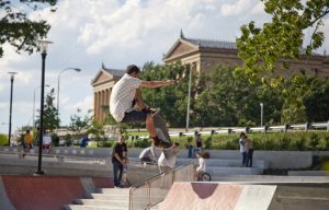 A color photograph of a man executing an aerial stunt on a skateboard using a ramp made of brick and concrete. The Philadelphia Museum of Art is visible behind him.