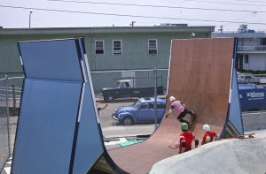 A color photograph of a skateboarder riding in a tall half-pipe. Two others stand by watching.