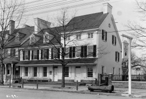 A black and white photograph of the Indian King Tavern, a white two-story building with a three-story attachment. The second floor has a row of windows with black shutters. A wooden horse trough is on the sidewalk in front.