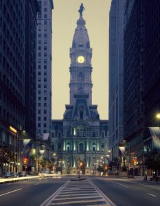 A color photograph of Philadelphia city Hall from South broad Street looking north. The building has a prominent tower with an illuminated clock face, topped with a dome and a statue of William Penn.