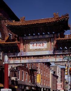 A color photograph of the Friendship Gate, a colorful traditional Chinese gateway ornamented with golden dragons. Chinese language characters are painted in red on a white background. They translate to read the words "Philadelphia Chinatown".