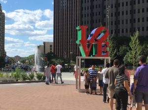 A color photograph of the LOVE Statue at John F. Kennedy Plaza, Philadelphia. A crowd waits in line for their turn to take a photo in front of the statue. There is a fountain behind it.