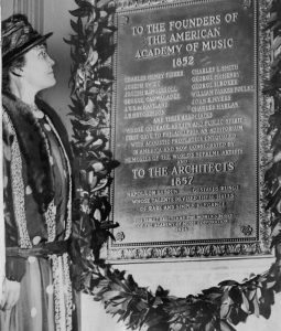 A black and white photograph of Frances Wister standing before a plaque celebrating the founders and architects of the Academy of Music. A wreath of evergreen leaves