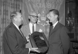 A black and white photograph of three men in suits. The two men on the left are presenting a plaque to Edmund Bacon, who stands to the right of them.