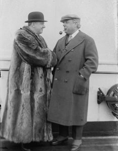 A black and white photograph of W. C. Fields, dressed in a full-length fur coat and bowler cap, shaking hands with Philip Goodman, who wears a long coat and cap and smokes a pipe. They stand on the deck of a ship.