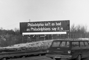 A black and white photograph of a billboard reading 'Philadelphia isn't as bad as philadelphians say it is." The billboard stands next to a highway. 1960s era vehicles pass by on the highway.