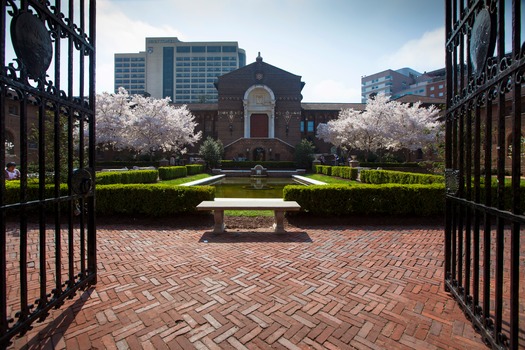 A color photograph of the main entrance of the University of Pennsylvania Museum. The photograph is taken through the iron gates and shows a formal garden with hedge rows and a reflecting pond with a stone bench in the foreground. Cherry trees in bloom flanking the front door.
