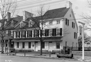 A black and white photograph of the Indian King Tavern in Haddonfield, a white Colonial-era three-story tavern with black window shutters. A wooden horse trough stands on the street in front. a hanging sign in the foreground reads "The Indian King".