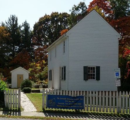 A color photograph of the Peter Mott House, a small white two-story house dating to the early nineteenth century. A white picket fence surrounds the grounds. A blue banner hangs on the fence marking the site as the home of the Lawnside historical society.