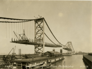 a black and white photograph of the unfinished Ben Franklin bridge taken from the Philadelphia side of the Delaware River. The cables and pillars are completed but there are large gaps in the road bed. In the foreground are several marinas and piers on the river. In the background is the skyline of the city of Camden.