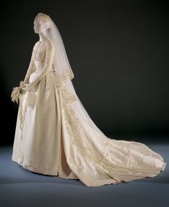 Mannequin wearing white wedding dress with lace details, a wreath-shaped crown, and matching clutch.