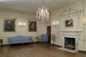 Interior of eighteenth-century parlor room with portraits on the walls, blue furniture, a fireplace, and a crystal chandelier.