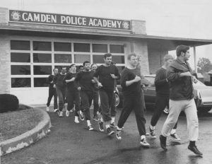 A black and white photograph of a line of men jogging on pavement in front of the Camden Police Academy. All of the men wear identical sweat suits except the instructor