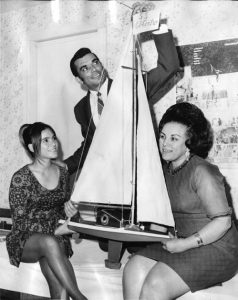 Black and white photograph showing two seated women holding a model boat. A Man stands behind them.