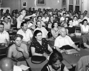 Black and white photograph showing adult students sitting at desks. Instructor is pointing at something in the front of the room.