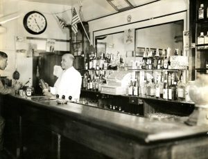 Black and white photograph depicting a man behind a bar preparing a drink while talking to a seated man.