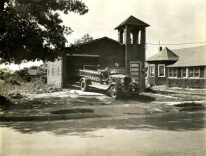 Black and white photograph depicting a man driving a fire engine out of a garage.