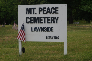 Color photograph depicting white sign that reads "Mount Peace Cemetery Lawnside Established 1900." An American flag is next to the sign. Graves are visible in the background.