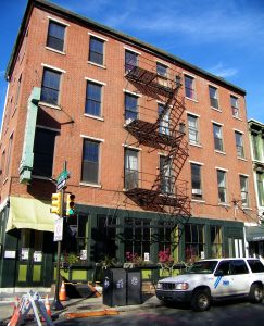 Color photograph showing a brick building with a simple iron fire escape. The ground level of the building has a storefront with many plants.