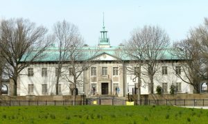 A color photograph of the American Swedish Historical museum, a large white building with a green cooper roof and cupola. The museum is surrounded by a black fence and stands in a grassy field surrounded by trees.