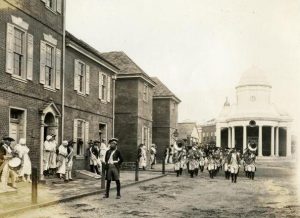 A black and white photograph showing a reconstruction of market street, Philadelphia, as it may have appeared during the American Revolution. Several two-story brick homes line an unpaved street. At the end of the street is a Greek Revival style building topped by a round dome. A marching band in Revolutionary War-style costumes marches down the street while men and women in colonial costumes stand in the doorways and on the sidewalks observing.