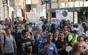 Color photograph showing a crowd of protesters, some holding signs, some with instruments, and some chanting.