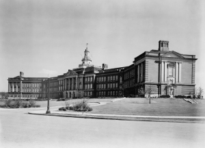 A black and white photograph of a large school building with greek-style columns at the front entrance and a tall white cupola on top.