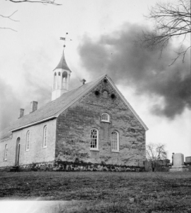 a black and white photograph of a small stone church with arched windows. The roof is topped by a white steeple with a weather vane. There is a small graveyard next do it. The photograph is taken in winter and several leafless trees stand in the background.