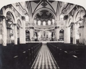 A black and white photo of the interior of the St. Joseph's Church in South Camden, New Jersey. The photograph is taken from the rear of the church down the central aisle, showing rows of pews and arched galleries along the walls. At the front of the church is an alter adorned with religious statues, housed in a domed alcove with an ornately detailed ceiling.