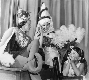 Black and white photograph of young women in fantasy costumes doing make up while a child looks on.