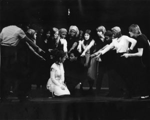 Black and white photograph showing a group of actors huddled around and a seated couple and pointing at them.