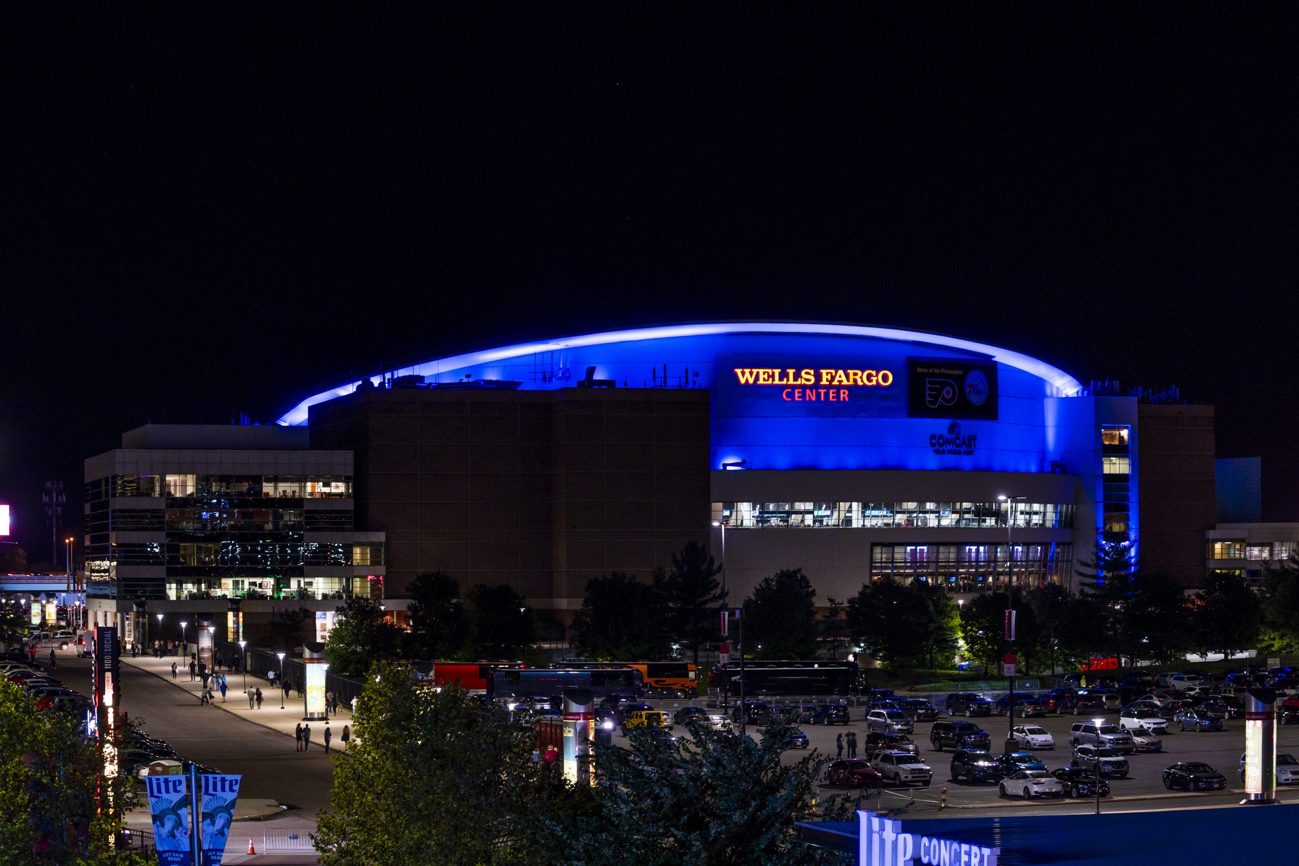 Color photograph depicting the Wells Fargo Center at night. The stadium is illuminated with blue lights and parking lot is in foreground.