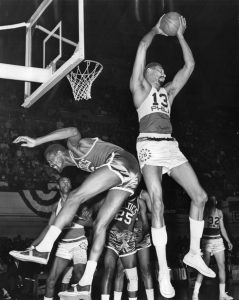 Black and white photograph showing Wilt Chamberlain colliding into a player on the opposing team as he jumps to take a shot.