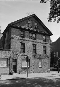 A black and white photograph of the Thaddeus Kosciuszko house. The three story brick home is in a state of disrepair with boarded windows, crumbling bricks, and evidence of previous repairs. A historical marker stands in front of it.