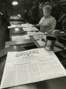 A black and white photograph of a newspaper printing room. A woman stands reading a paper set on a desk. In the foreground, a folded newspaper reads "GWIAZDA. Polish Star. Final issue of Gwiazda."