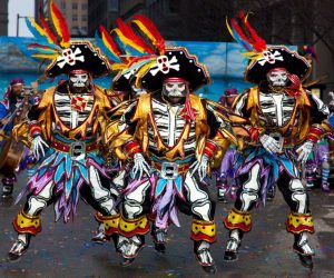 A color photograph of members of the Polish-American string Band performing in the 2011 mummer's parade. Four male dancers wear pirate-inspired skeleton costumes of brightly colored fabrics, skull masks, and hats with colorful feathers. Behind them, a row of musicians in similar costumes play musical instruments. Colorful confetti litters the ground.