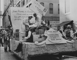 A black and white photograph of the 1952 Pulaski Day Parade. Adults wearing a doctor and nurse costume stand on a float featuring a hospital bed. A child lays in the bed with bandages on her head and other children sit on boxes labeled "medical supplies," "penicillin," and "vaccines". In the background, a giant letter and envelope give a plea for medical supplies.