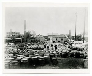 Black and white photograph depicting men standing on a dock completely covered in barrels full of sugar.