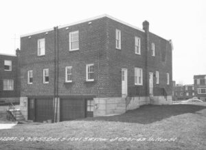 A black and white photo of the rear of a two-story brick twin house with garage doors.