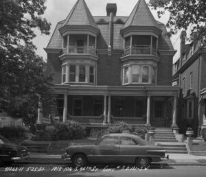 A black and white photo of twin houses built in the Queen Anne style. They feature a large porch held up by columns and a prominent bay window and balcony topped by a turret.