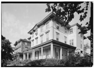 A black and white photo of two twin three-story Italianate houses at Woodland Terrace. One is faced in white stucco, the other in dark stone. Both have porches along the front and sides of the ground floor. Maple trees line the street.