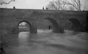 A black and white photograph of a stone arched bridge crossing a creek in winter. Two people stand on the bridge looking over the water.