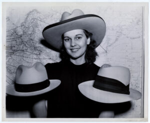 A black and white photograph of a woman in a light-colored cowboy hat with a dark brim. She holds a light-colored hat with a dark band in each hand. Behind her is a map of the world with shipping routes labeled on it.