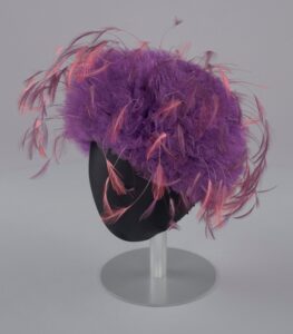 A color photograph of a women's hat on a black hat stand against a white background. The hat is made of ruffled purple net fabric and adorned with long pink feathers