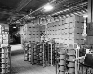 A black and white photograph of men's hats on racks in the Stetson factory. A row of stacked wooden crates embossed with the stetson logo stands in the background. A woman is placing more crates into the row.