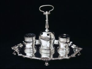 A color photograph of a silver inkstand. The round silver plate has four short feet keeping it raised slightly above the surface it sits on. On it sit three silver ink pots, the center one with a large handle protruding from the top.
